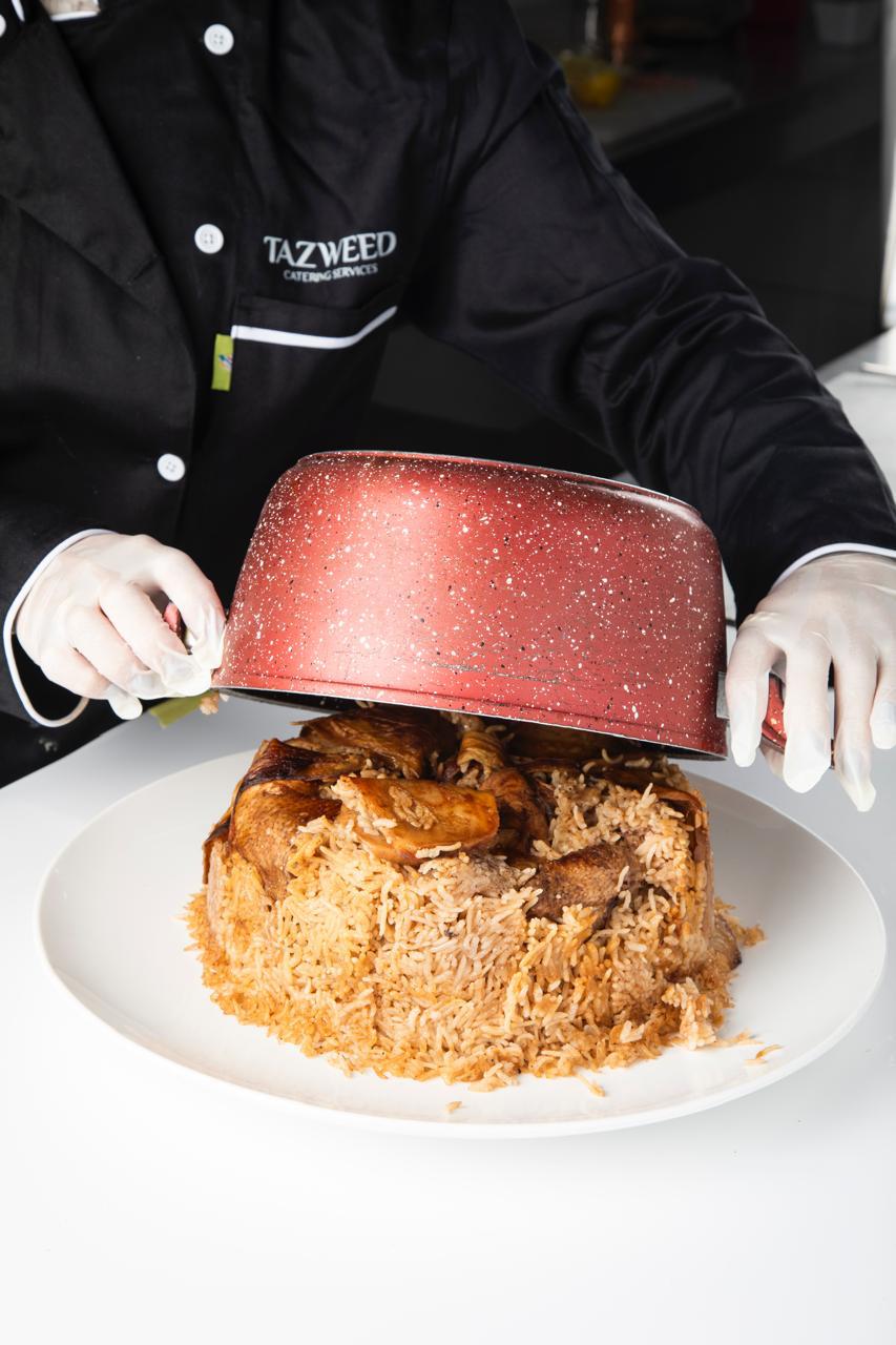 Person in a black Tazweed chef's coat lifting the lid of a dish on a white plate.