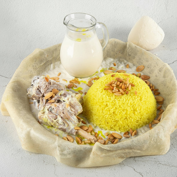 Mansaf dish with yellow rice and meat, served in a bread bowl with a glass of milk, on a light gray background. Tazweed Catering Services branding at the bottom.